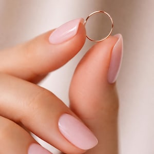 K&ouml;nnte beinhalten: Ein zarter, d&uuml;nner Ring aus Ros&eacute;gold, der zwischen zwei Fingern mit hellrosa Nagellack gehalten wird. Der Ring hat ein schlichtes, minimalistisches Design, ideal f&uuml;r den t&auml;glichen Gebrauch. Der Hintergrund ist eine weiche, neutrale Farbe.