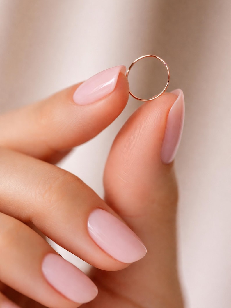 K&ouml;nnte beinhalten: Ein zarter, d&uuml;nner Ring aus Ros&eacute;gold, der zwischen zwei Fingern mit hellrosa Nagellack gehalten wird. Der Ring hat ein schlichtes, minimalistisches Design, ideal f&uuml;r den t&auml;glichen Gebrauch. Der Hintergrund ist eine weiche, neutrale Farbe.