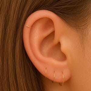 May include: Close-up of an ear adorned with delicate gold hoop earrings. Three small hoops are in the earlobe, and a single hoop is in the upper ear. The earrings have a simple, minimalist design.