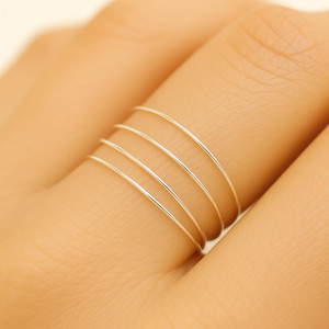 May include: A hand wearing a set of four thin, silver rings. The rings are simple bands, each a separate ring, stacked on a finger. The rings are made of a shiny, metallic material, reflecting light. The background is a soft, neutral color.
