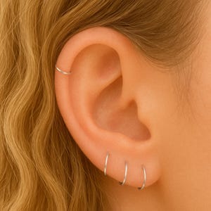 May include: Close-up of an ear with four small, silver hoop earrings. The earrings are simple and minimalist, with a thin, circular design. The ear is set against a backdrop of wavy, blonde hair.