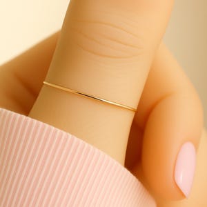 May include: A delicate, thin gold ring worn on a finger. The ring is a simple band, and the finger is adorned with a light pink nail polish. The person is wearing a light pink long-sleeved shirt.