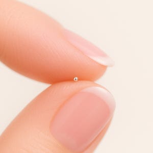 May include: A tiny, silver-colored sphere held between two fingers. The sphere appears to be a small bead or ball, possibly jewelry or a component of a larger item. The image is a close-up, highlighting the size and detail of the object.
