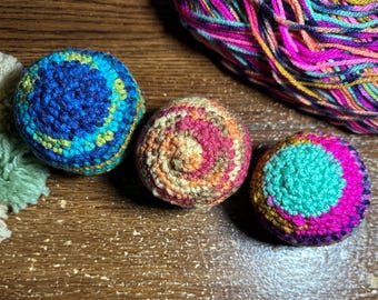 May include: Three spherical, crocheted items arranged in a row. The left sphere features blue, green, and yellow yarn. The center sphere displays a spiral pattern of pink, orange, and tan. The right sphere shows a mix of bright pink, teal, and yellow.