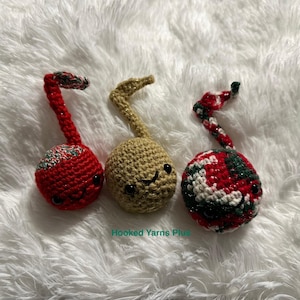 May include: Three crocheted musical note ornaments in red, green, and beige. Each ornament has a smiling face and is decorated with a red and green plaid pattern.