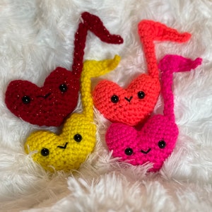 May include: Four crocheted musical notes in shades of red, yellow, orange, and pink. Each note has a heart-shaped head with a smiling face and a stem. The notes are arranged on a fluffy white surface.