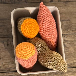 May include: A collection of handmade crochet vegetables, including a sweet potato, two acorn squash, and a carrot, in a cardboard container. The vegetables are in shades of orange, pink, and brown, with detailed stitching.