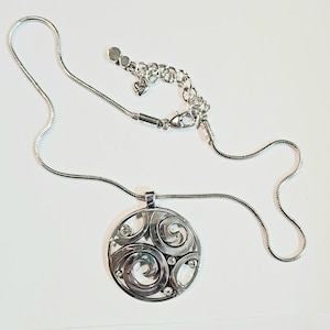 以下が含まれることがあります： 円形ペンダントが付いたシルバーカラーのネックレス。ペンダントは、渦巻き模様と小さな透明なクリスタルをあしらったオープンワークデザインです。チェーンは細いスネークチェーンスタイルで、長さは調節可能です。