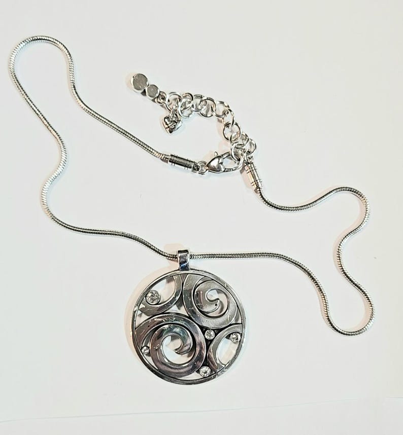 以下が含まれることがあります： 円形ペンダントが付いたシルバーカラーのネックレス。ペンダントは、渦巻き模様と小さな透明なクリスタルをあしらったオープンワークデザインです。チェーンは細いスネークチェーンスタイルで、長さは調節可能です。