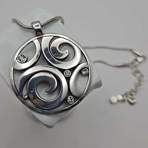 以下が含まれることがあります： 渦巻き模様と埋め込まれたクリアクリスタルが特徴の、円形デザインのシルバーペンダントネックレス。 ネックレスはスネークチェーンと調節可能な留め金が付いています。 ペンダントの直径は約5cmです。