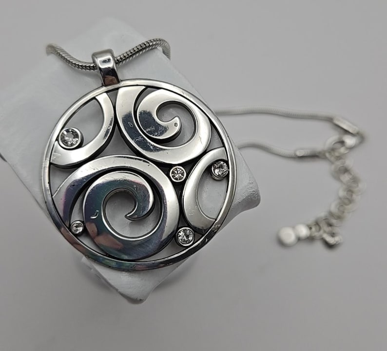 以下が含まれることがあります： 渦巻き模様と埋め込まれたクリアクリスタルが特徴の、円形デザインのシルバーペンダントネックレス。 ネックレスはスネークチェーンと調節可能な留め金が付いています。 ペンダントの直径は約5cmです。