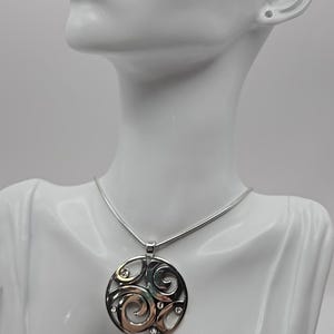 以下が含まれることがあります： 渦巻き模様と小さな透明な石をあしらった円形デザインのシルバーペンダントネックレス。ペンダントは細いシルバーチェーンから吊り下げられています。ネックレスは白いマネキンのバストに飾られています。