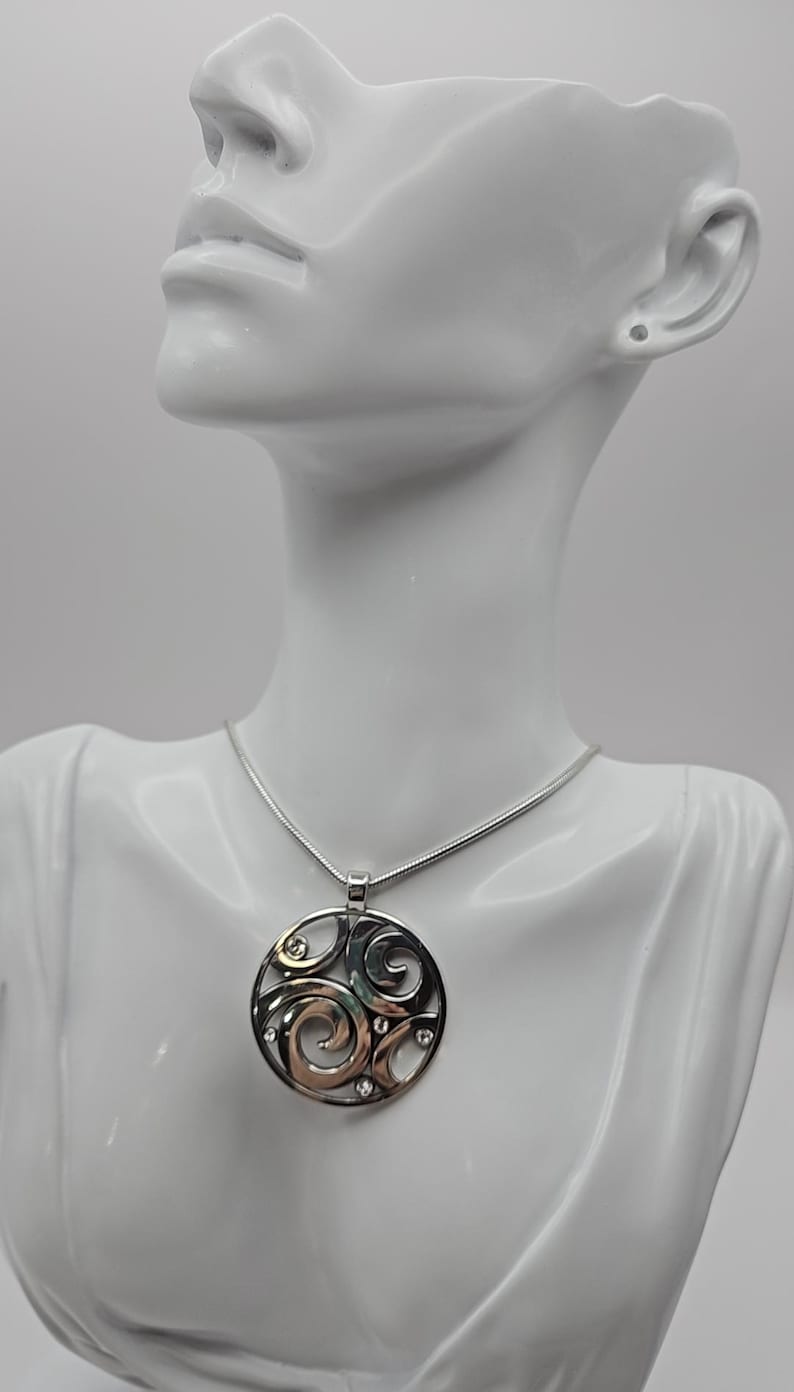 以下が含まれることがあります： 渦巻き模様と小さな透明な石をあしらった円形デザインのシルバーペンダントネックレス。ペンダントは細いシルバーチェーンから吊り下げられています。ネックレスは白いマネキンのバストに飾られています。