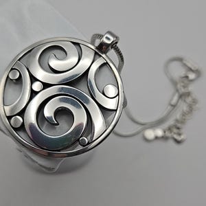 以下が含まれることがあります： 渦巻き模様と小さな円形アクセントが特徴の、円形デザインのシルバーペンダントネックレス。ペンダントは繊細なシルバーチェーンに付いています。ネックレスは白い表面に表示されています。