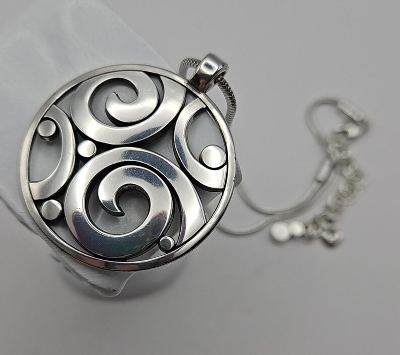 以下が含まれることがあります： 渦巻き模様と小さな円形アクセントが特徴の、円形デザインのシルバーペンダントネックレス。ペンダントは繊細なシルバーチェーンに付いています。ネックレスは白い表面に表示されています。