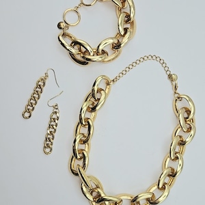 May include: A gold-toned jewelry set featuring a chunky chain design. The set includes a bracelet with a toggle clasp, a necklace, and a pair of dangle earrings. The jewelry is displayed on a white surface.
