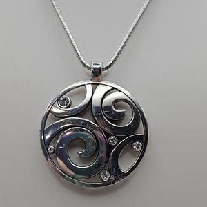 以下が含まれることがあります： シルバーカラーのペンダントネックレス。渦巻き模様のオープンワークデザインの円形ペンダント。ペンダントには小さなクリアクリスタルが装飾されています。ネックレスは繊細なチェーンです。