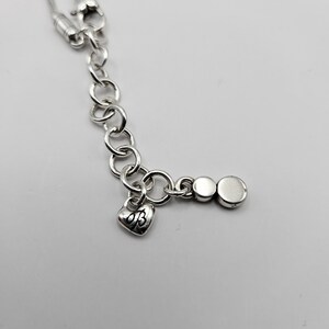 以下が含まれることがあります： ハート型のチャームと丸いエンドピースが付いた、シルバーカラーのネックレスチェーン。ハートチャームには「AB」のイニシャルが刻印されています。チェーンは、小さく相互接続されたリンクでできています。