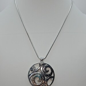 以下が含まれることがあります： 円形デザインのシルバーペンダントネックレス。ペンダントは、渦巻く形と小さな透明なクリスタルからなるオープンワークパターンが特徴です。ネックレスは白いマネキンに展示されています。