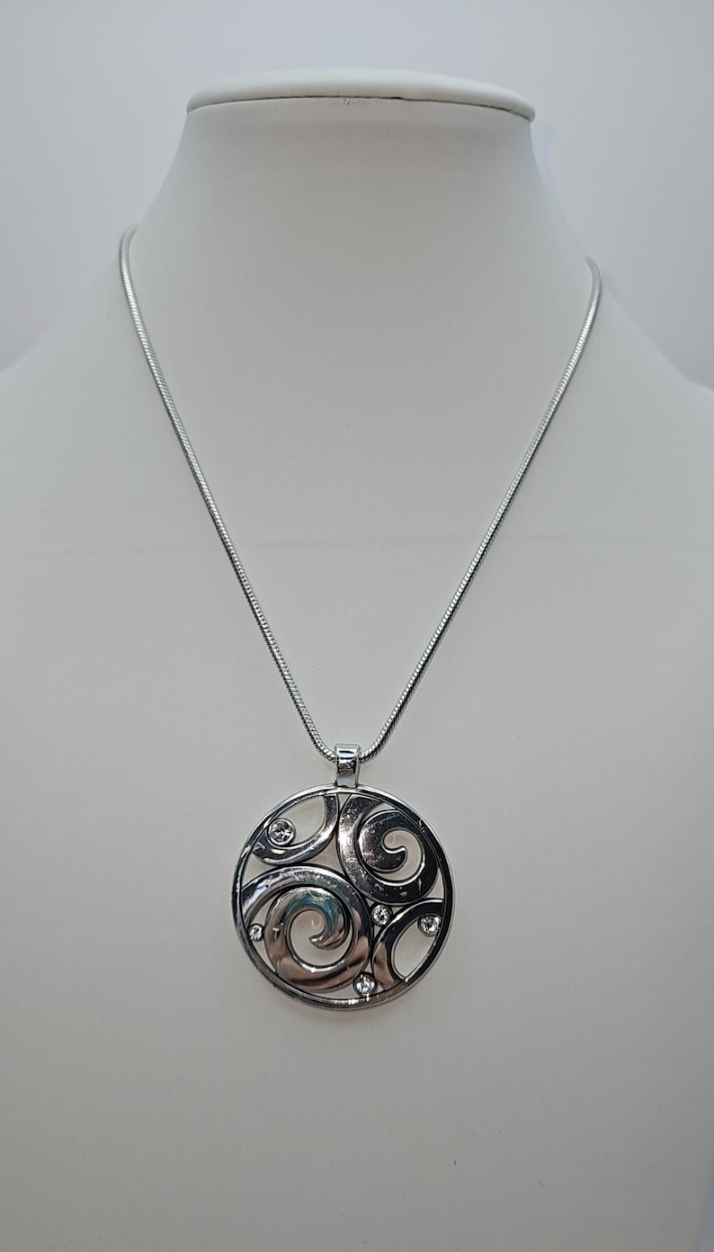 以下が含まれることがあります： 円形デザインのシルバーペンダントネックレス。ペンダントは、渦巻く形と小さな透明なクリスタルからなるオープンワークパターンが特徴です。ネックレスは白いマネキンに展示されています。