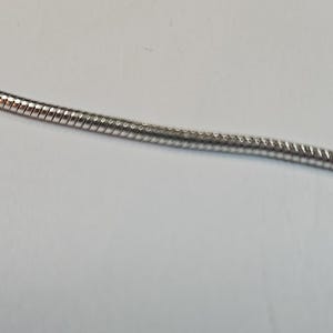 以下が含まれることがあります： 銀色のスネークチェーンネックレスのクローズアップ。チェーンは、きつく織られたテクスチャードデザインです。背景はプレーンな白い表面です。ネックレスはジュエリーアイテムです。