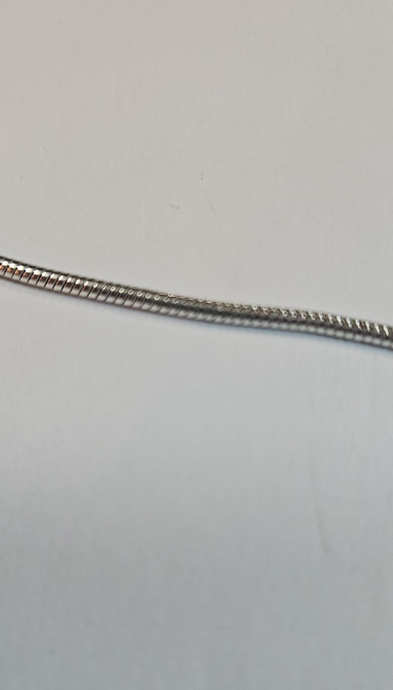 以下が含まれることがあります： 銀色のスネークチェーンネックレスのクローズアップ。チェーンは、きつく織られたテクスチャードデザインです。背景はプレーンな白い表面です。ネックレスはジュエリーアイテムです。