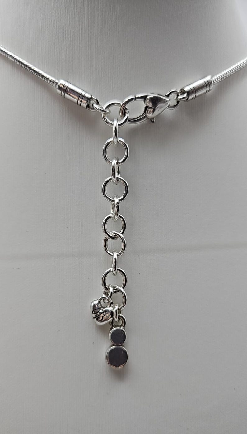 以下が含まれることがあります： シルバーカラーのネックレス、スネークチェーンのデザイン。ロブスタークラスプ留めと、ハート型のチャームと2つの丸いチャームが付いた調節可能なチェーンが特徴です。白い背景に展示されています。