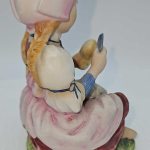 May include: A porcelain figurine of a seated girl wearing a pink and white dress and bonnet. The girl has blonde braids and is holding a small object. The figurine is painted with soft colors and sits on a green base.