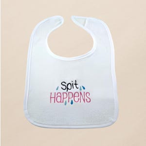 May include: White baby bib with the embroidered text "Spit Happens" in black and pink. The bib has a curved neckline and a soft, absorbent texture, designed to catch spills and drool.