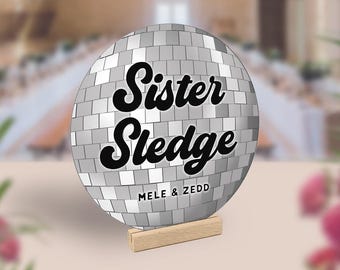 May include: A silver disco ball with the text "Sister Sledge" in black lettering and "Mele & Zedd" in smaller black lettering below. The disco ball is on a wooden stand.