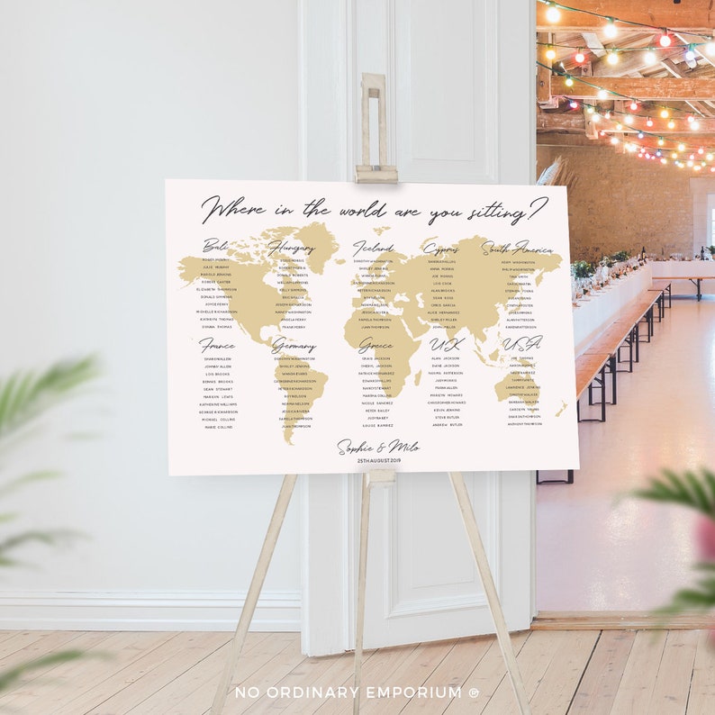 Op de afbeelding: Een gouden wereldkaart zitplaatskaart met de tekst "Where in the world are you sitting?" bovenaan. De kaart is verdeeld in secties voor verschillende landen, waaronder Frankrijk, Duitsland, Griekenland, VK, VS, Bali, Hongarije, Ierland, Cyprus en Zuid-Amerika. De namen van de gasten staan onder elk land vermeld. De kaart staat op een houten ezel met de tekst "Sophia & Milo" en de datum "23rd August 2019" onderaan.