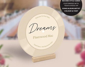 May include: A cream-colored record-style card with the word "Dreams" and "Fleetwood Mac" printed on it. The card is displayed on a wooden stand. Text on the card reads "Welcome To Your Table" and "Lisa & Harry - 15th September 2024". A black circle displays "Record Style Cards".