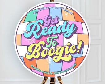 Puede incluir: Un letrero de boda redondo con el texto "Get Ready To Boogie!" en una fuente retro. El letrero tiene un diseño de bola de discoteca colorido con colores pastel. El letrero está sobre un caballete de madera.
