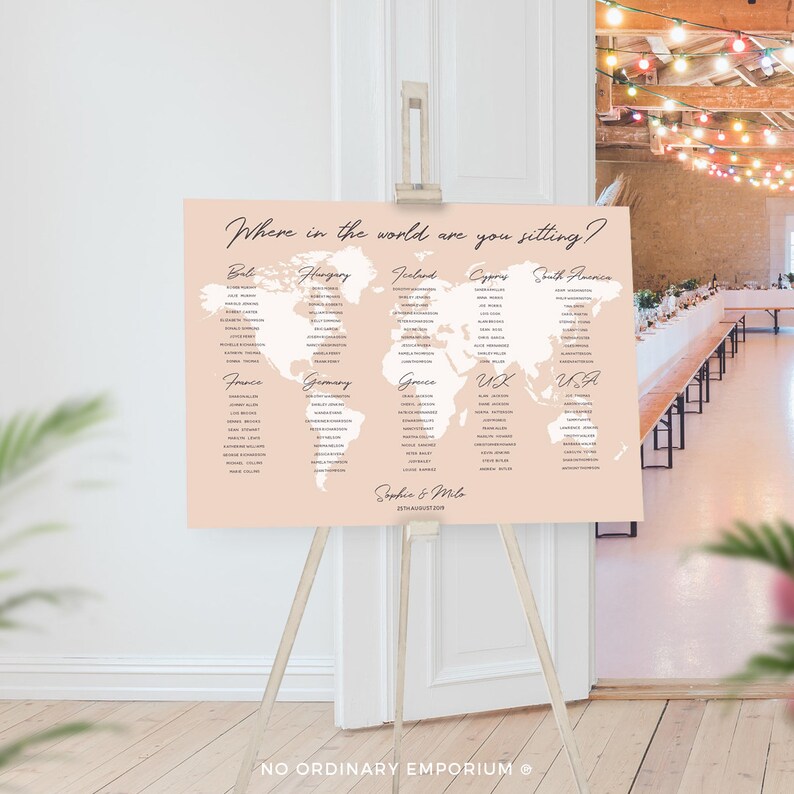 Op de afbeelding: Een roze en witte wereldkaart zitplaatskaart met de tekst "Waar in de wereld zit je?" bovenaan. De kaart is verdeeld in secties voor verschillende landen, waaronder Bali, Hongarije, IJsland, Cyprus, Zuid-Amerika, Frankrijk, Duitsland, Griekenland, VK en de VS. De namen van de gasten staan ​​onder elk land vermeld. De kaart staat op een houten ezel en zegt "Sophia & Miles" en "27 augustus 2018" onderaan.
