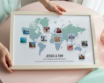 May include: A framed world map print with a light wood frame. The map is light blue and features small photos marking travel locations. The text "JESSIE & TOM - OUR WORLD ADVENTURES" is displayed at the bottom, with additional text below.
