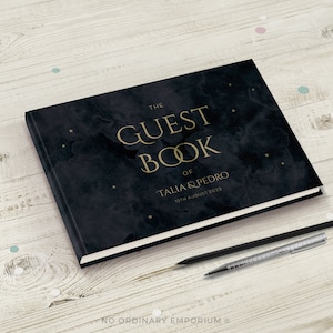 May include: A black guest book with gold lettering that reads "The Guest Book of Talia & Pedro 15th August 2023". The cover has a dark blue watercolor background with gold stars.