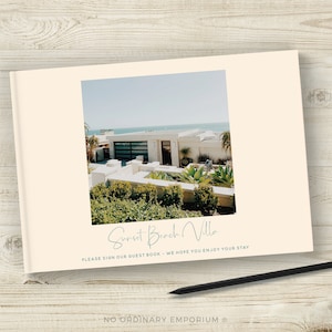 May include: A guest book with a photo of a white house with a blue roof and a view of the ocean. The text on the cover reads "Sunset Beach Villa" and "Please sign our guest book - We hope you enjoy your stay".