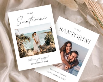 May include: Two white table cards with the word "Santorini" in elegant script. Each card features a photograph of a couple and the text "Where we got engaged 20th March 2022". The cards are on a soft, neutral-toned surface.