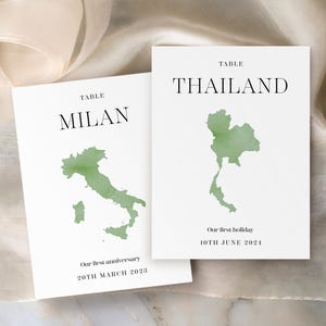 May include: Two white cards with the word "TABLE" at the top. One card says "MILAN" with a green watercolor Italy map and "Our first anniversary 20TH MARCH 2023". The other says "THAILAND" with a green watercolor Thailand map and "Our first holiday 10TH JUNE 2024".