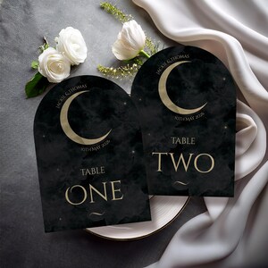 May include: Two black and gold wedding table cards with an arched top. Each card features a gold crescent moon, the names "JACKIE & THOMAS", and the date "10TH MAY 2026". The cards are labeled "TABLE ONE" and "TABLE TWO".