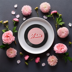 May include: A black vinyl record with a pink label that says "Bride" in a retro font. The record is on a white plate surrounded by pink flowers.