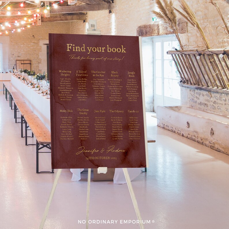 May include: A burgundy wedding seating chart with gold lettering. The chart is titled "Find your book" and lists guests' names under different book titles. The chart is on a wooden easel and the text at the bottom reads "Jennifer & Andrea 14th October 2023".