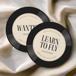 May include: Two black vinyl record coasters with beige centers. One says "WANTED" and the other says "LEARN TO FLY" with "FOO FIGHTERS" below. Both have "WELCOME TO YOUR TABLE" at the top and names and a date at the bottom.