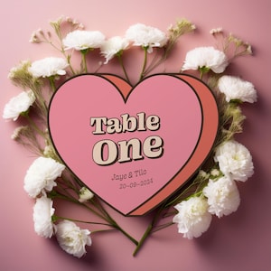 May include: A pink heart-shaped table number with the text "Table One" in a retro font. The text "Jaye & Tilo 20-08-2024" is below the table number. The heart is surrounded by white flowers on a pink background.