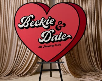 May include: A large, red heart-shaped sign with the names "Beckie & Dale" in white script, and the date "5th January 2024". The sign has a black outline and is displayed on a black easel. The color set is red, black, and white.