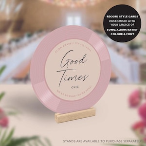 May include: A pink record-style card with the words "Good Times" in elegant script. The card includes the names "Alisa & Dave" and the date "7th July 2024". The card is displayed on a small wooden stand. Text on the card reads "CHIC" and "WE'RE SO GLAD YOU'RE HERE!"