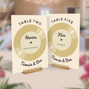 May include: Two wedding table cards with a vintage vinyl record design. One card says "Heroes" by David Bowie, the other says "Kiss" by Prince. Both cards have the names "Tamsin & Ben" at the bottom, and are held by wooden stands.