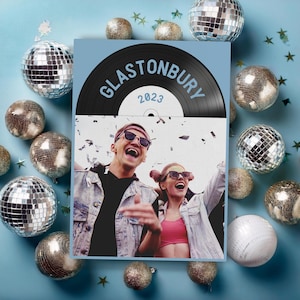 May include: A promotional poster for Glastonbury 2023, featuring a vinyl record design with the festival name and year. The poster showcases a photo of two people wearing sunglasses and smiling, surrounded by disco balls and star-shaped confetti on a blue background.