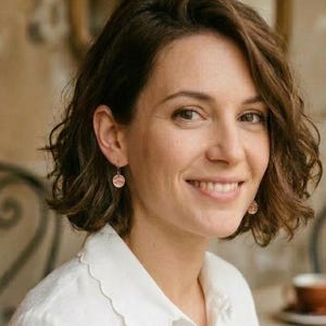 May include: A woman with short, wavy brown hair smiles, wearing a white collared shirt and small round earrings. The background shows a cafe setting with tables, chairs, and a mirror.