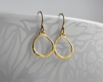 Puede incluir: Un par de pendientes colgantes de color dorado con una forma simple y orgánica. Los pendientes están hechos de metal y tienen un acabado liso.